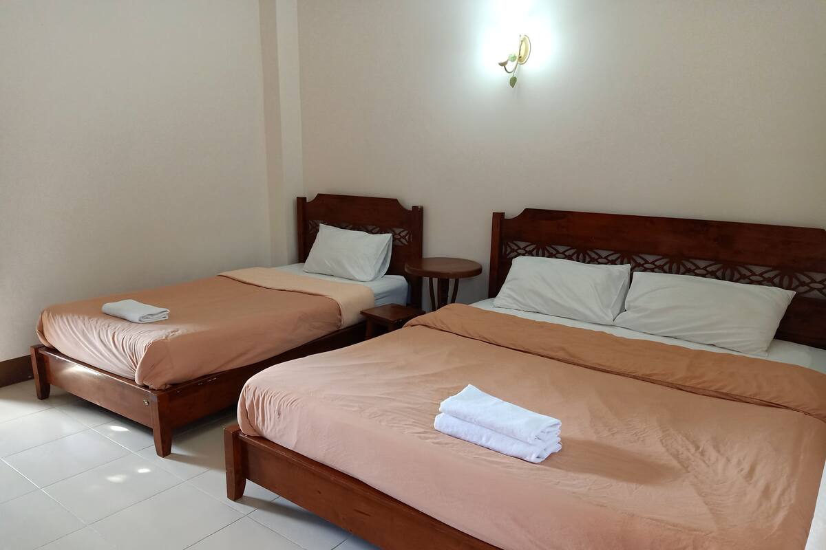 A hotel room with two wooden beds, each with a white pillow and a folded white towel. There is a small round table between the beds, and a wall-mounted light fixture above it.
