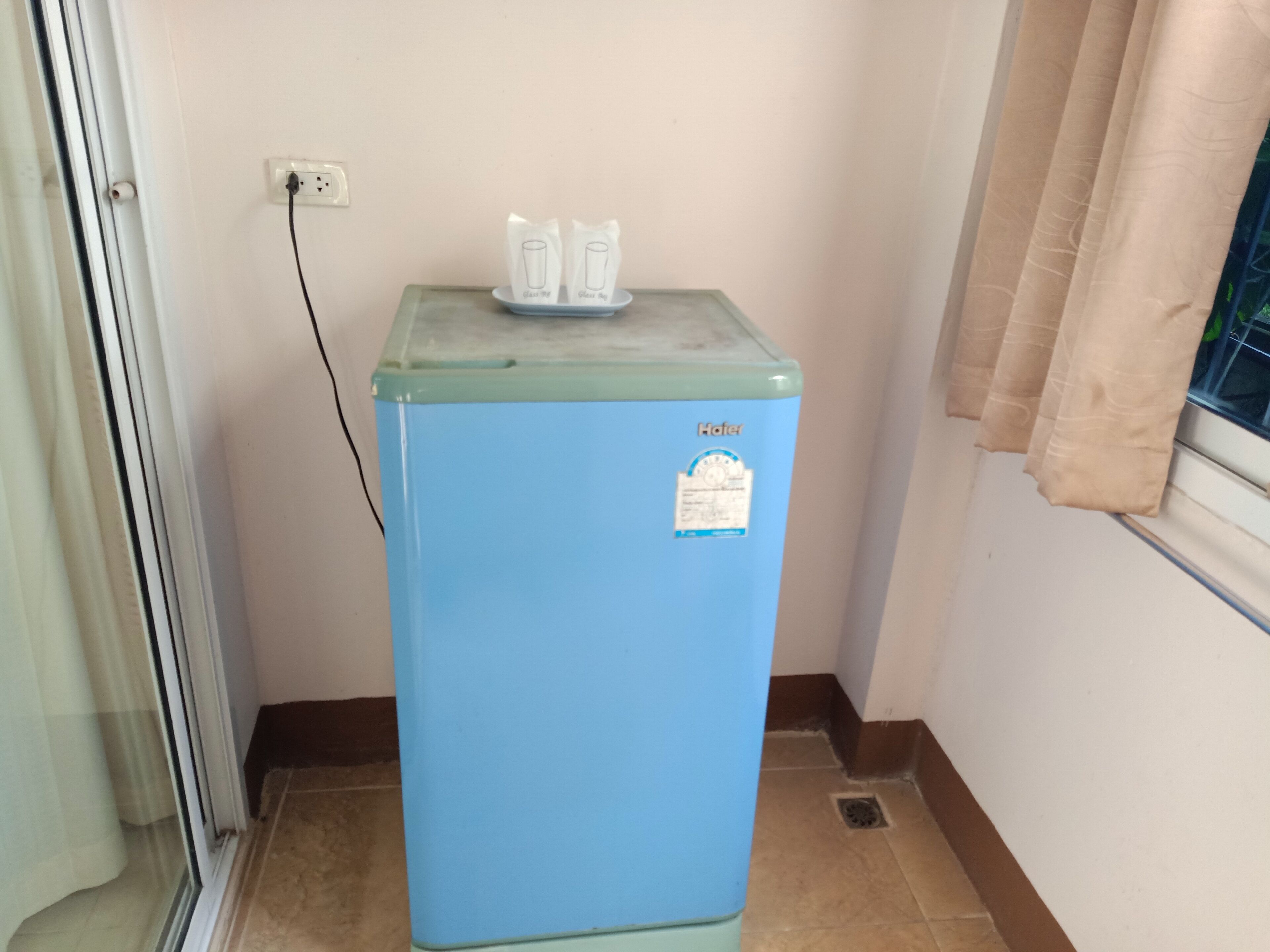 Un refrigerador azul con una etiqueta adhesiva, un enchufe y una ventana con cortinas.