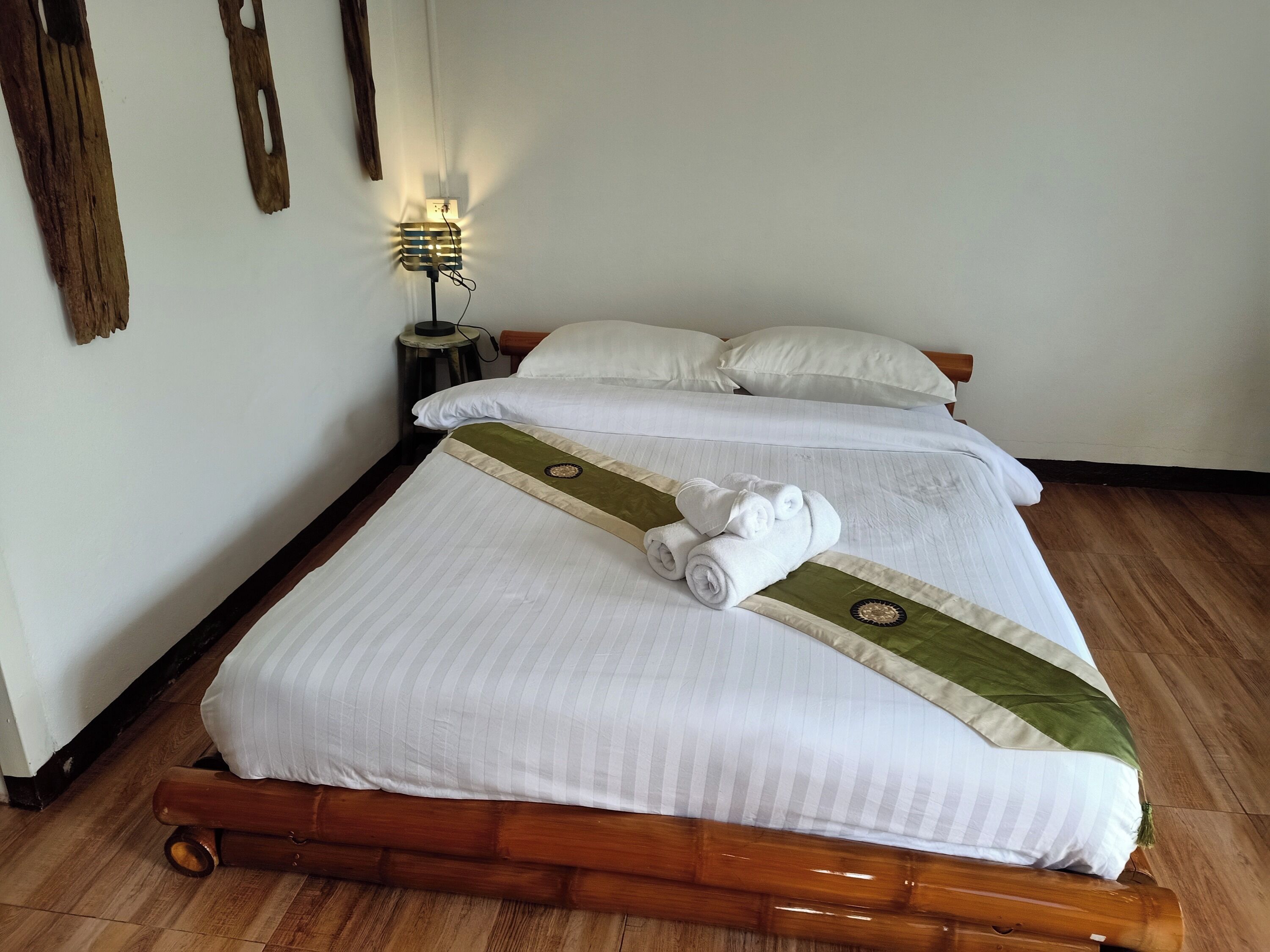 Una cama bien tendida con sábanas blancas y un corredor de cama verde y beige, una mesita de noche pequeña con una lámpara, y un suelo de madera.