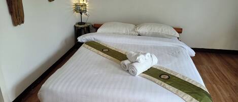 Una cama bien tendida con sábanas blancas y un corredor de cama verde y beige, una mesita de noche pequeña con una lámpara, y un suelo de madera.