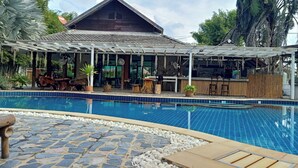 A swimming pool with a stone border, a covered bar area, and a building with a tiled roof in the background. - The Forest Hill Cafe & Homestay (San Sai)