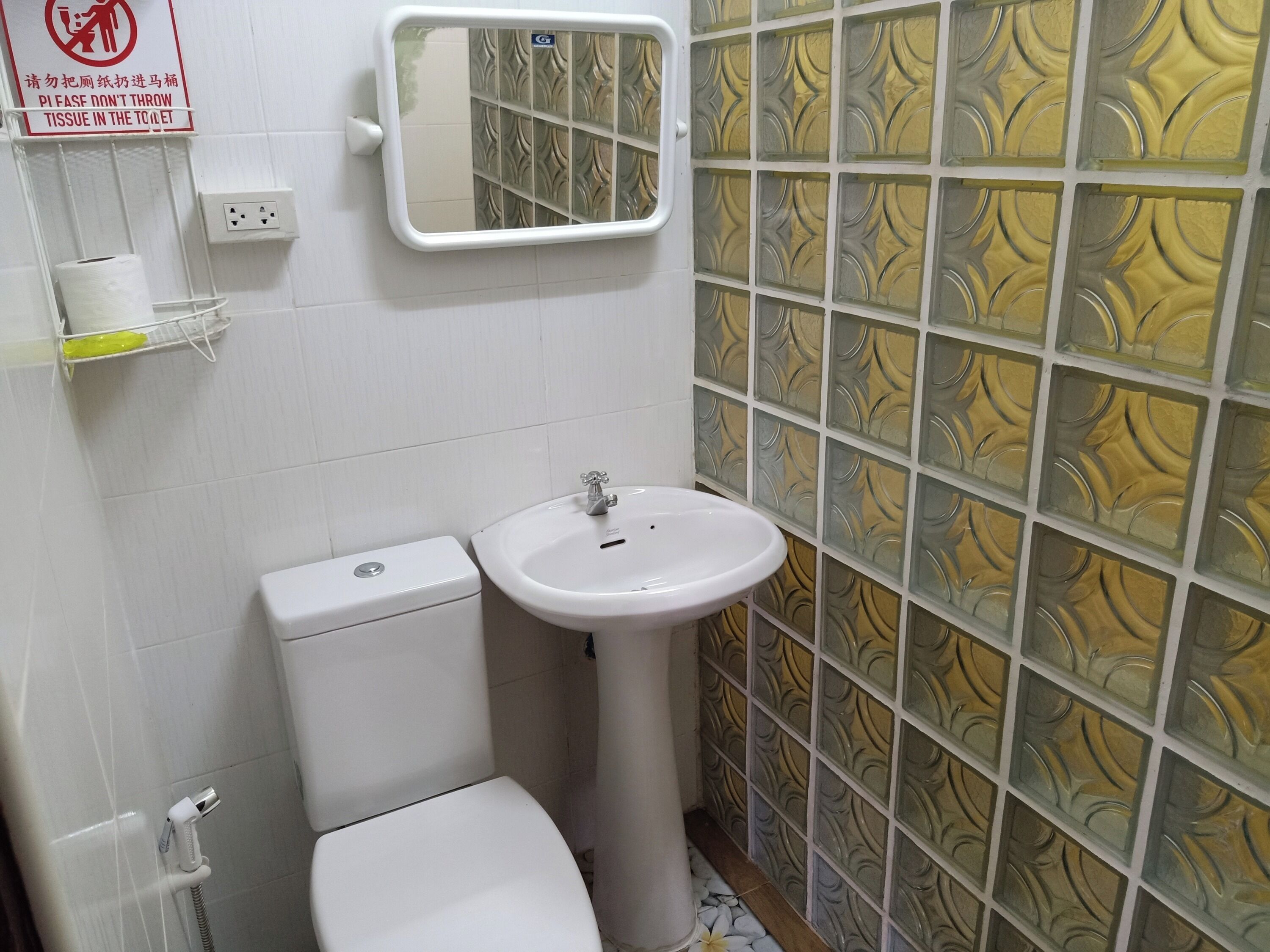 Egy fürdőszoba, amelyben WC, mosdókagyló és tükör található. A WC felett egy tábla, a falon pedig egy konnektor van.