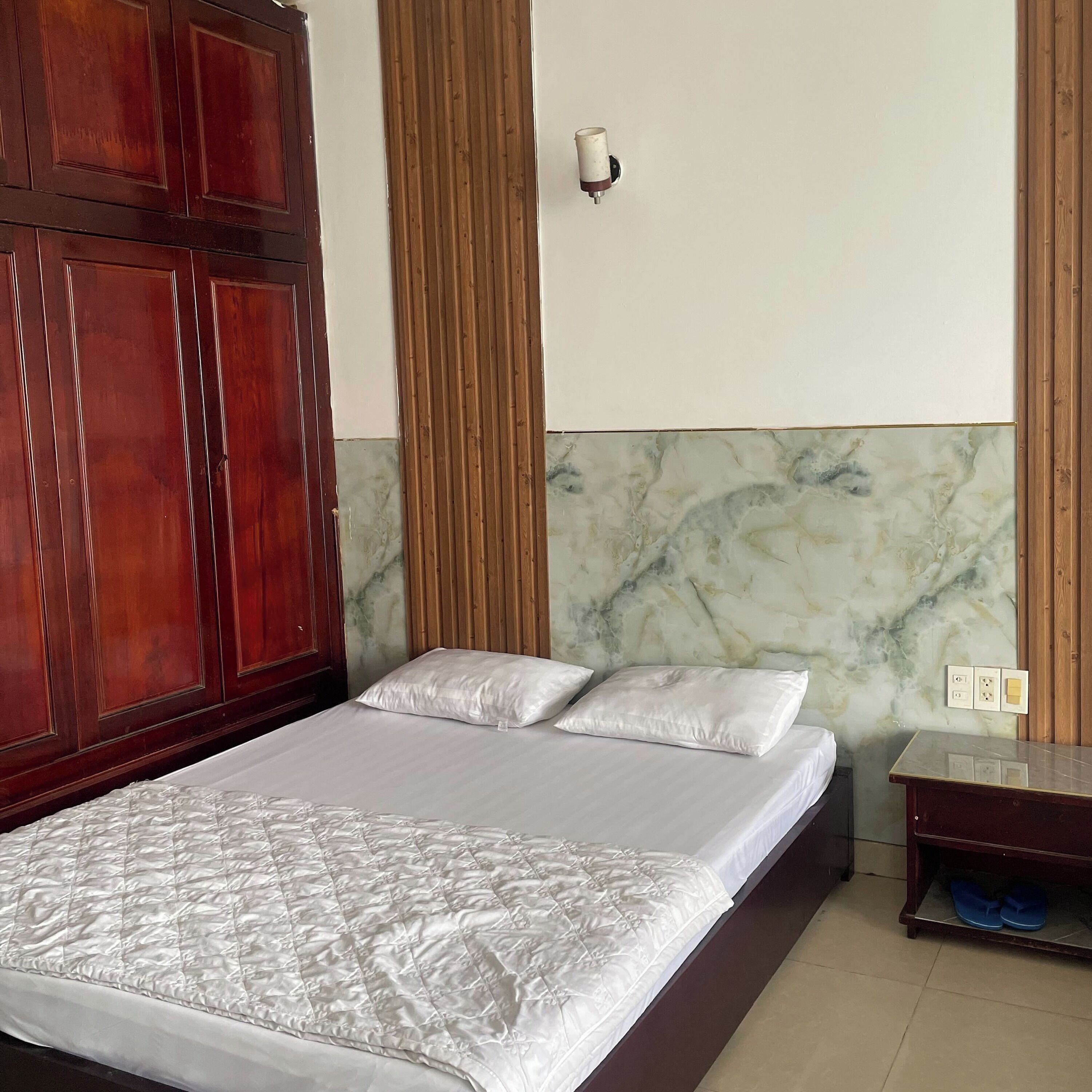A neatly made bed with a quilted white cover, a wooden wardrobe, a marble-patterned wall, and a small bedside table with a lamp.