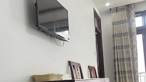 Ein Zimmer mit einem an der Wand befestigten Flachbildfernseher, einer Holztür, einem gemusterten Vorhang und einem kleinen Tisch mit einem Korb und Bilderrahmen.