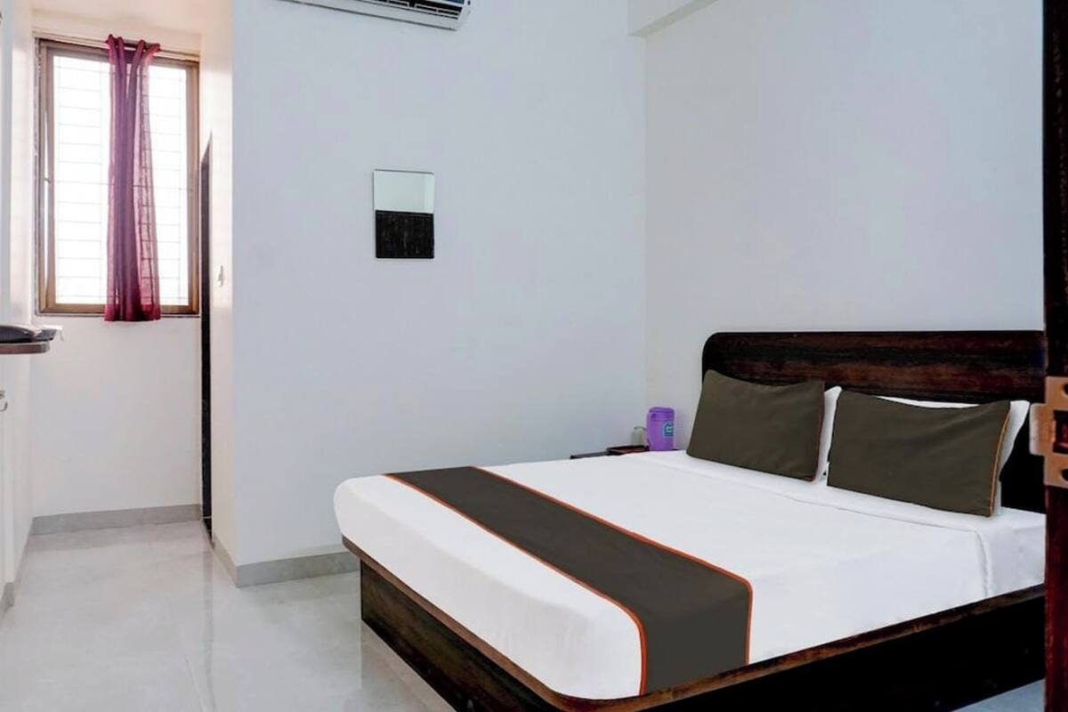 Een slaapkamer met een bed, een raam met gordijnen, een luchtverkoeler aan de muur en een tafeltje met een paars object.