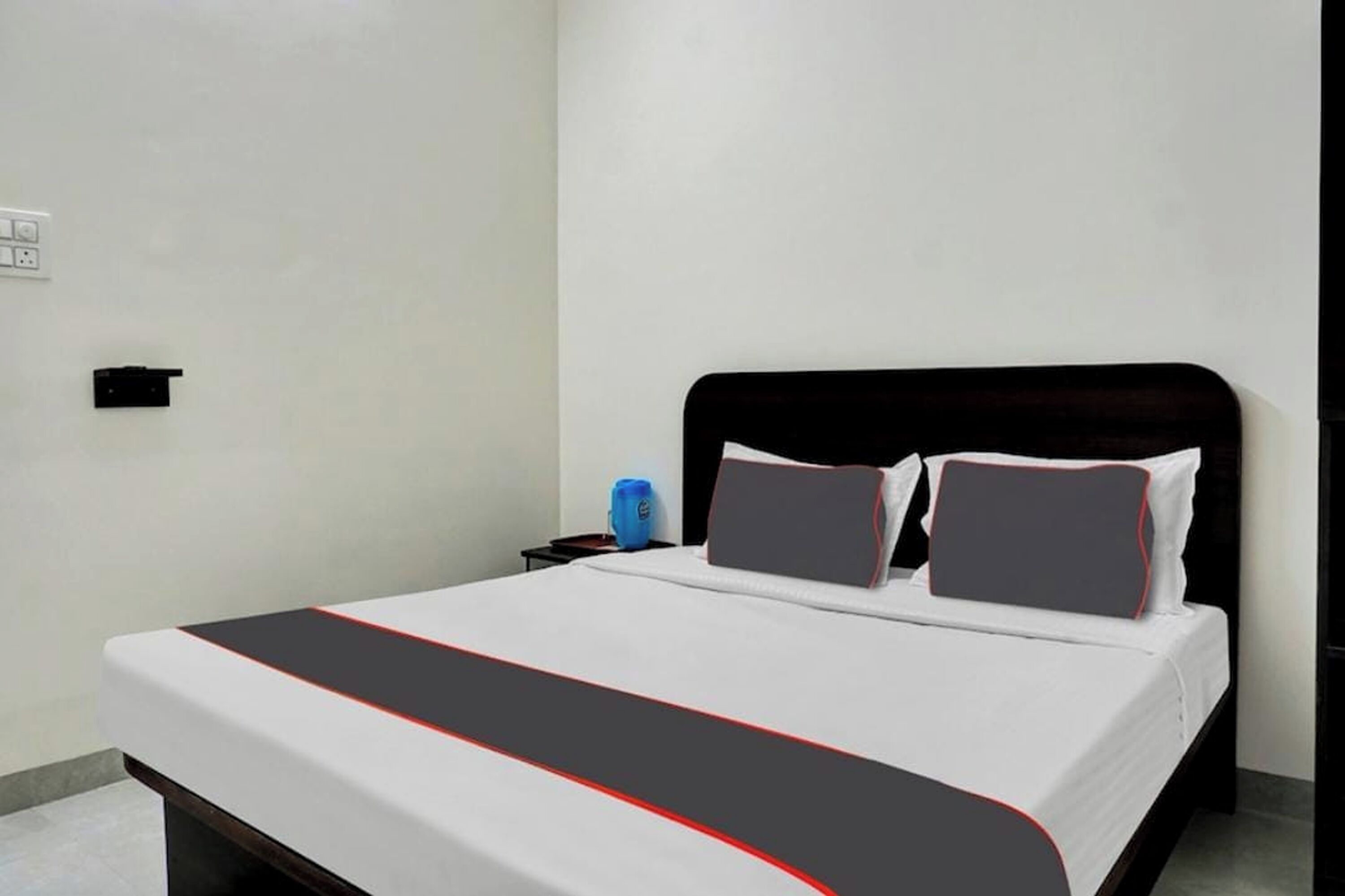 A neatly made bed with a black and red accent, a dark headboard, and a small bedside table with a blue object on it.