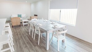 Een moderne eetruimte met een lange tafel voor zes personen, witte stoelen en een groot raam met zonwering.