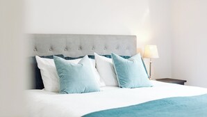 Een net opgemaakt bed met een grijs bekleden hoofdeinde, witte en lichtblauwe kussens, en een blauwgroene plaid.