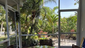 Une terrasse fermée par des baies vitrées, avec une table ronde et des chaises, entourée d’une végétation luxuriante et offrant une vue sur l’extérieur.