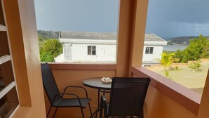 A small balcony with a tiled floor, two black chairs, a round table, and a view of a building and greenery.