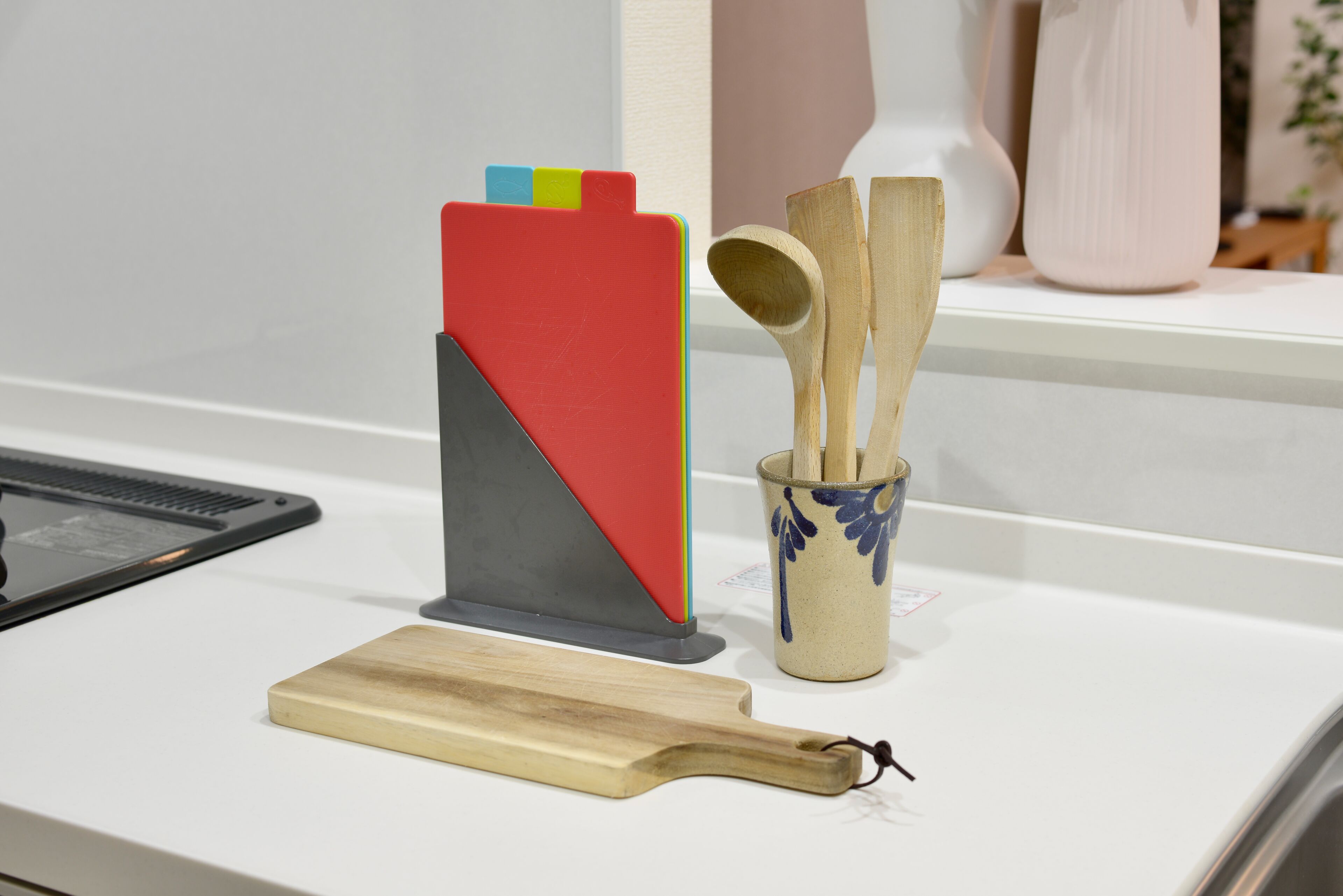 Una encimera de cocina con tabla de cortar, bloque de cuchillos rojo y gris, utensilios de madera y una taza de cerámica.