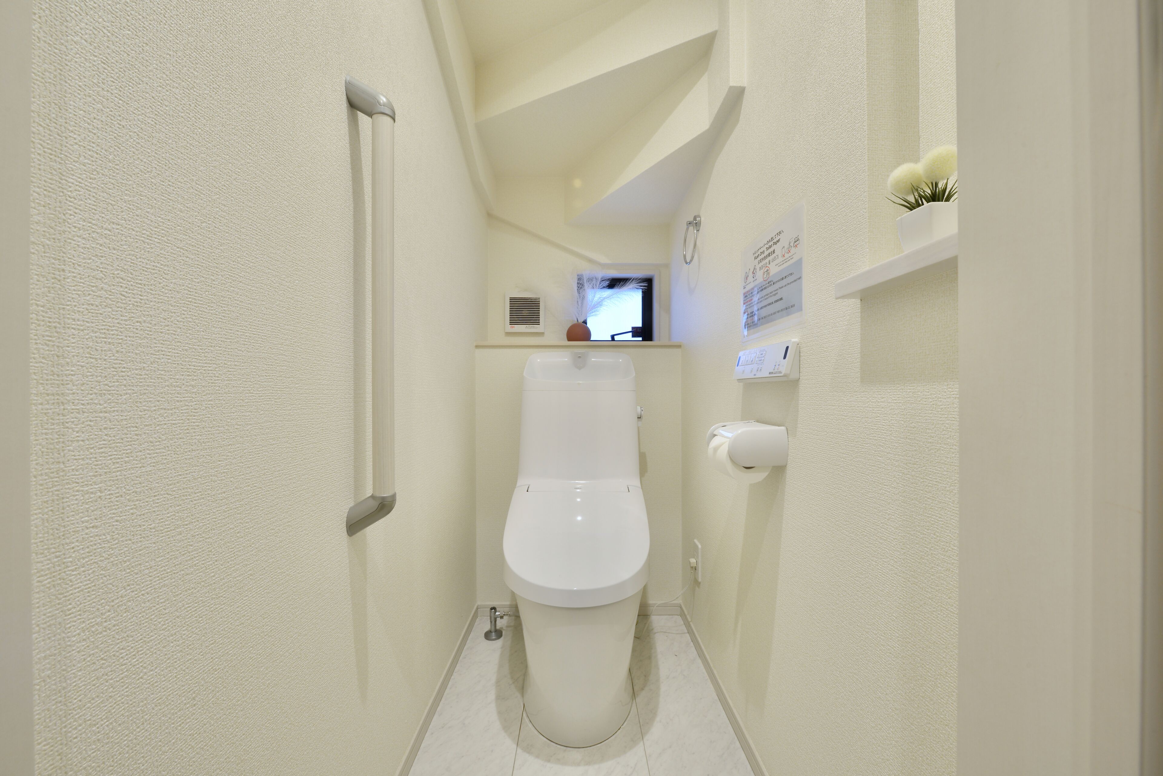 A small, narrow bathroom with a toilet, a handrail, a shelf with a potted plant, and a wall-mounted sign.