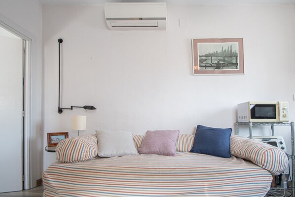 A bedroom with a bed, pillows, a lamp, an air conditioner, a framed picture, and a microwave.