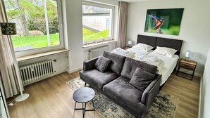 A modern bedroom with a large bed, a grey sofa, a small round table, and a view of the garden through the windows. - Rothaarsteig by Interhome (Winterberg)