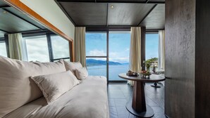 A modern hotel room with a large bed, a round table with a vase and fruit, and a view of the sea and mountains through large windows. - Dali Lanting Seaview Resort Hotel (Dali)