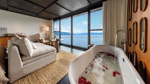 A modern hotel room with a large window, a bathtub with rose petals, a sofa, and a wooden wall panel. - Dali Lanting Seaview Resort Hotel (Dali)