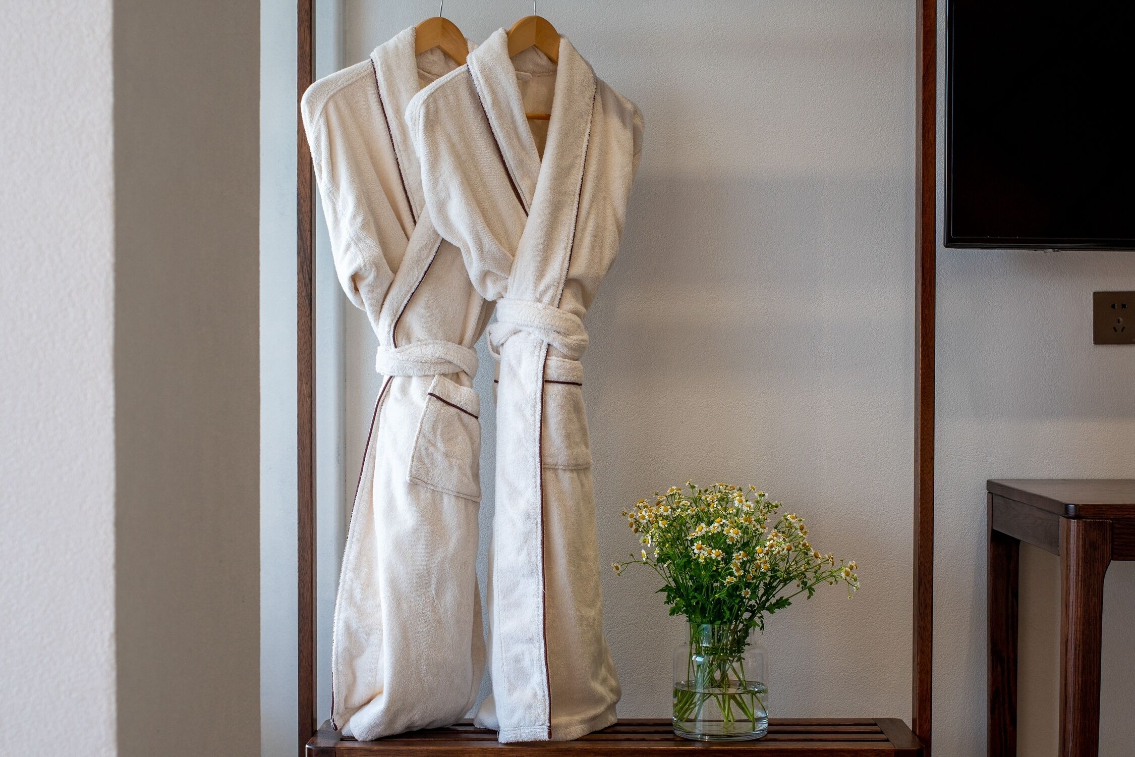 Dos albornoces blancos colgados en un perchero de madera, un pequeño jarrón con flores blancas y una mesa de madera oscura.