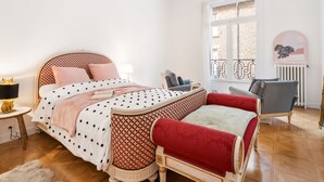 A bedroom with a bed, bedside table, lamp, chair, and a view of the outside.