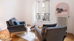 A living room with a grey armchair, a sofa, a white radiator, and a view of a balcony with a brick wall and a window.