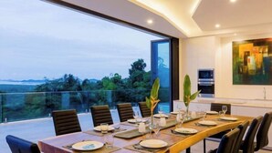 A dining area with a long table set for a meal, large windows offering a view of the outdoors, and modern kitchen appliances in the background. - Yamu Hills Panoramic Ocean View 6 Bed Luxury Vila (Pa Klok)