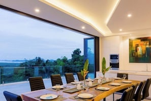 Un comedor con una mesa larga dispuesta para una comida, ventanas amplias con vistas al exterior y electrodomésticos modernos al fondo.