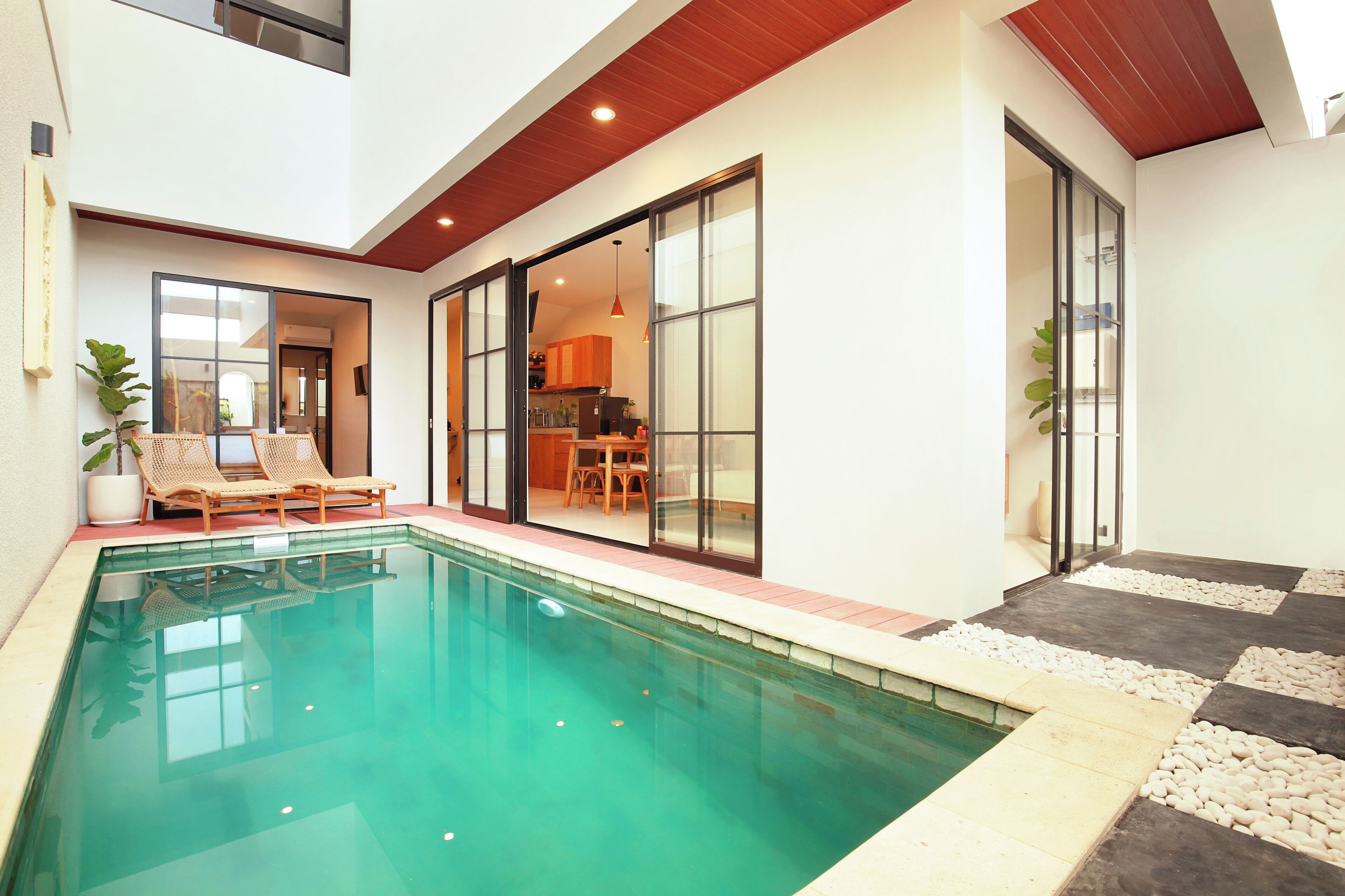 A modern indoor pool area with a clear pool, lounge chairs, and a dining area visible through glass doors.
