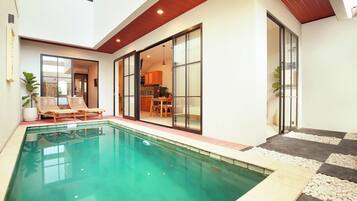 A modern indoor pool area with a clear pool, lounge chairs, and a dining area visible through glass doors.