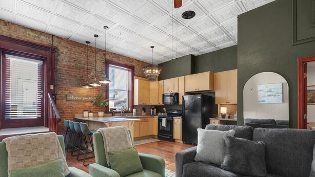A modern kitchen with a brick wall, wooden cabinets, and a black refrigerator. There are two green armchairs and a gray sofa in the living area.
