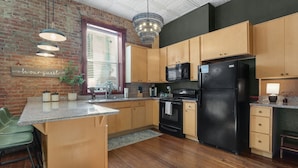 A kitchen with wooden cabinets, a black refrigerator, a microwave, and a stove. There is a window with blinds, a chandelier, and a sign that says "be our guest".