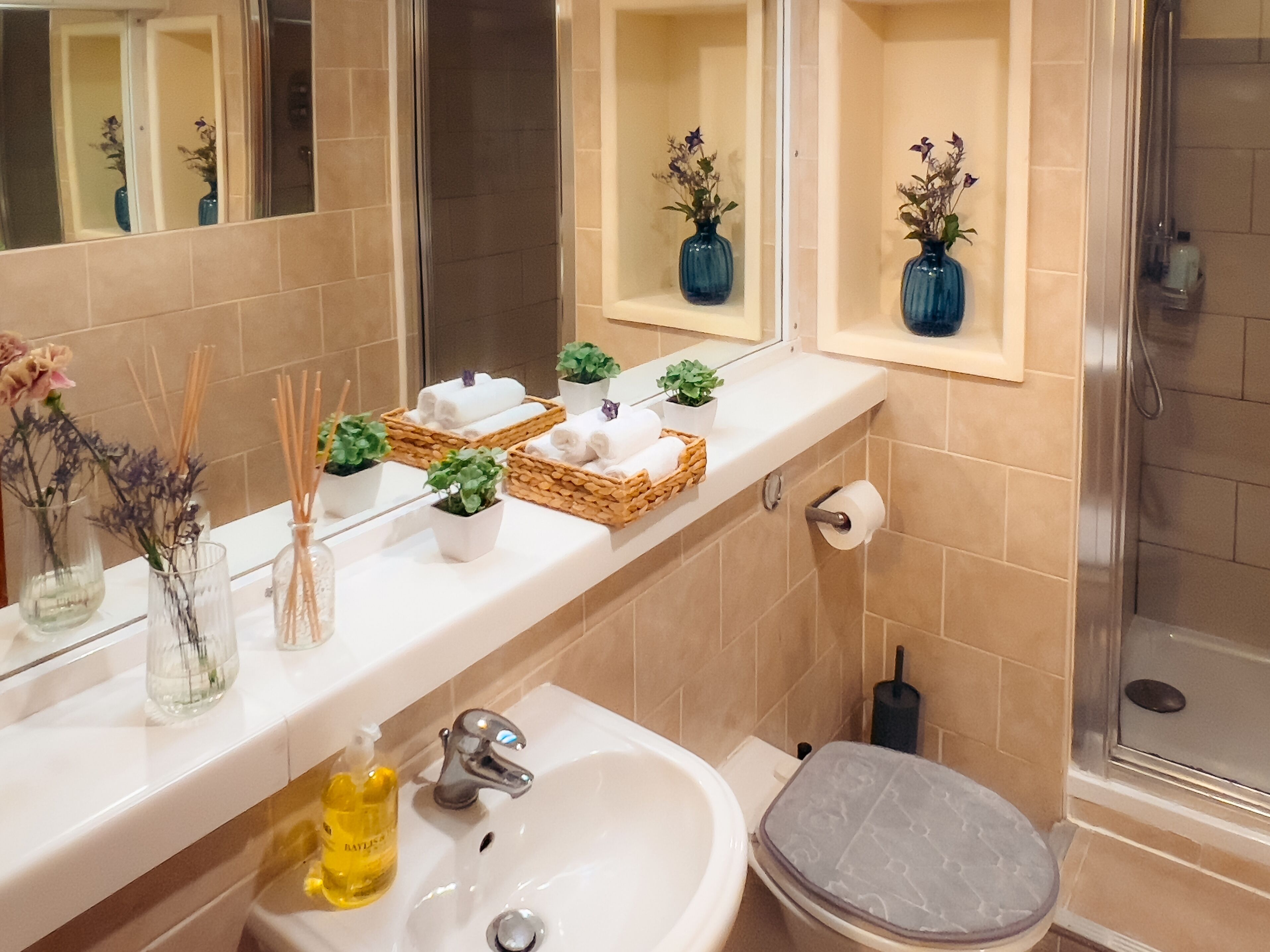 Une salle de bain avec un lavabo blanc, des toilettes et un espace douche. On y trouve deux vases bleus ornés de fleurs sur le meuble-lavabo, ainsi qu’une bouteille de lotion sur le lavabo.