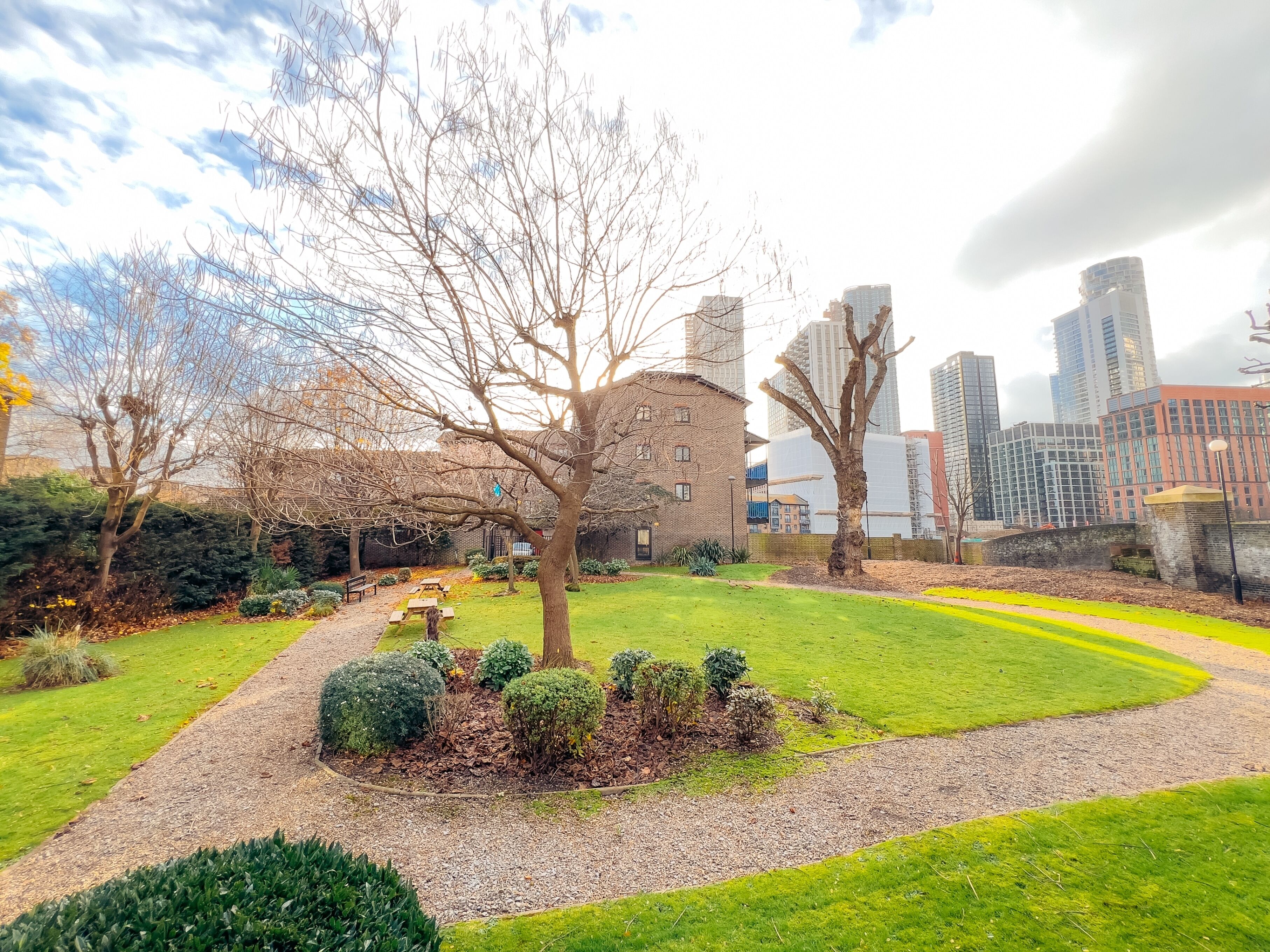 A park with a picnic table, a brick building, and modern high-rise buildings in the background.