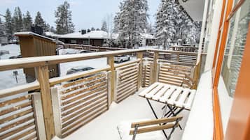 Een balkon met houten hekwerk, een witte, beklede bank en uitzicht op besneeuwde bomen en gebouwen.