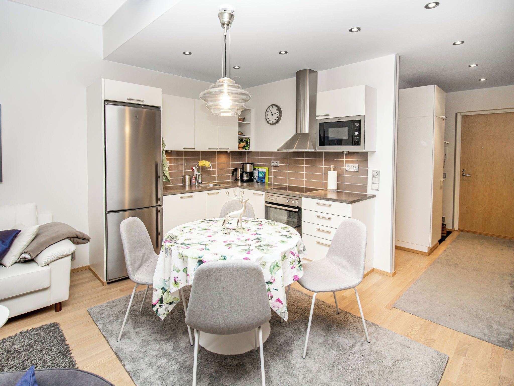Een moderne keuken met een koelkast, magnetron, fornuis en spoelbak. Er is een eettafel met stoelen en een klok aan de muur.
