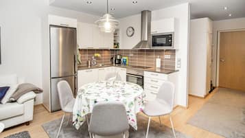 Een moderne keuken met een koelkast, magnetron, fornuis en spoelbak. Er is een eettafel met stoelen en een klok aan de muur.