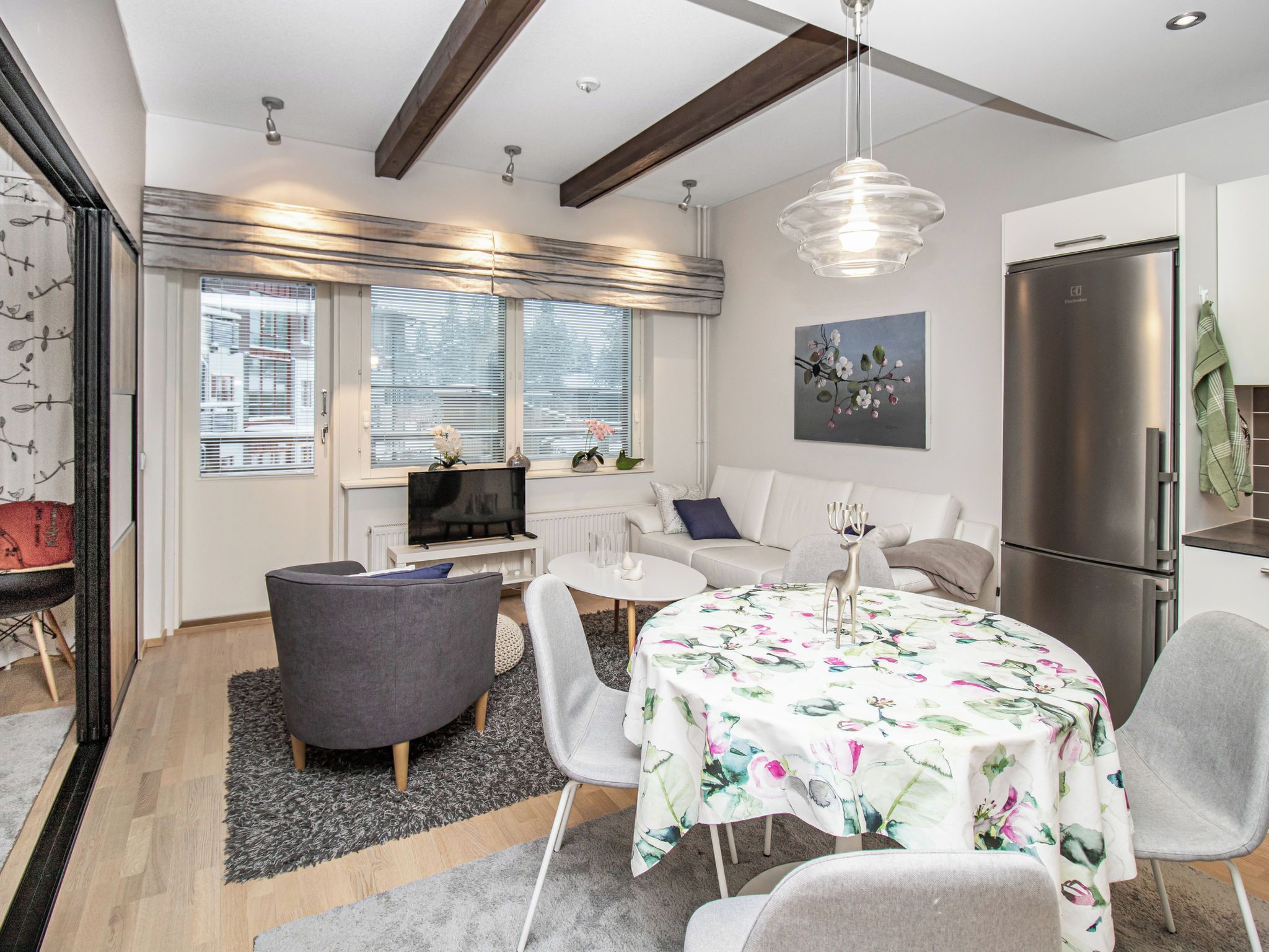 A modern living room with a sofa, a dining table with floral tablecloth, a grey armchair, and a kitchen area with a refrigerator.