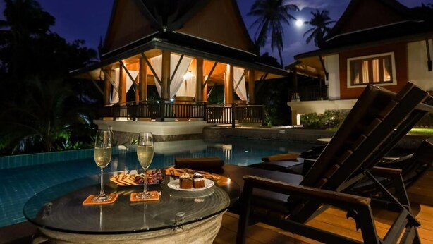 A poolside area with lounge chairs and a table set for dining, overlooking a building with a distinctive architectural style. - Eden Pool Villa  (Rawai)