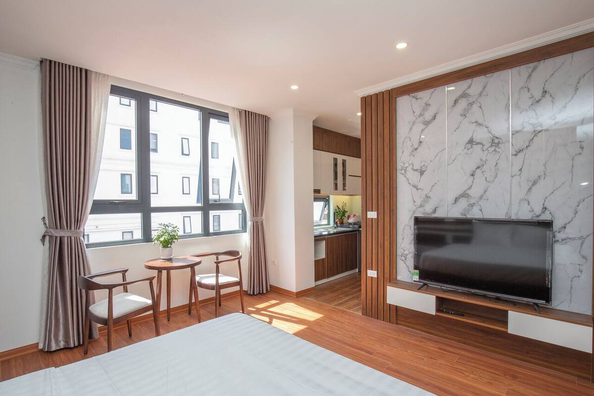 A modern hotel room with a large TV, wooden flooring, a dining area with a table and chairs, and a view of the city through large windows.