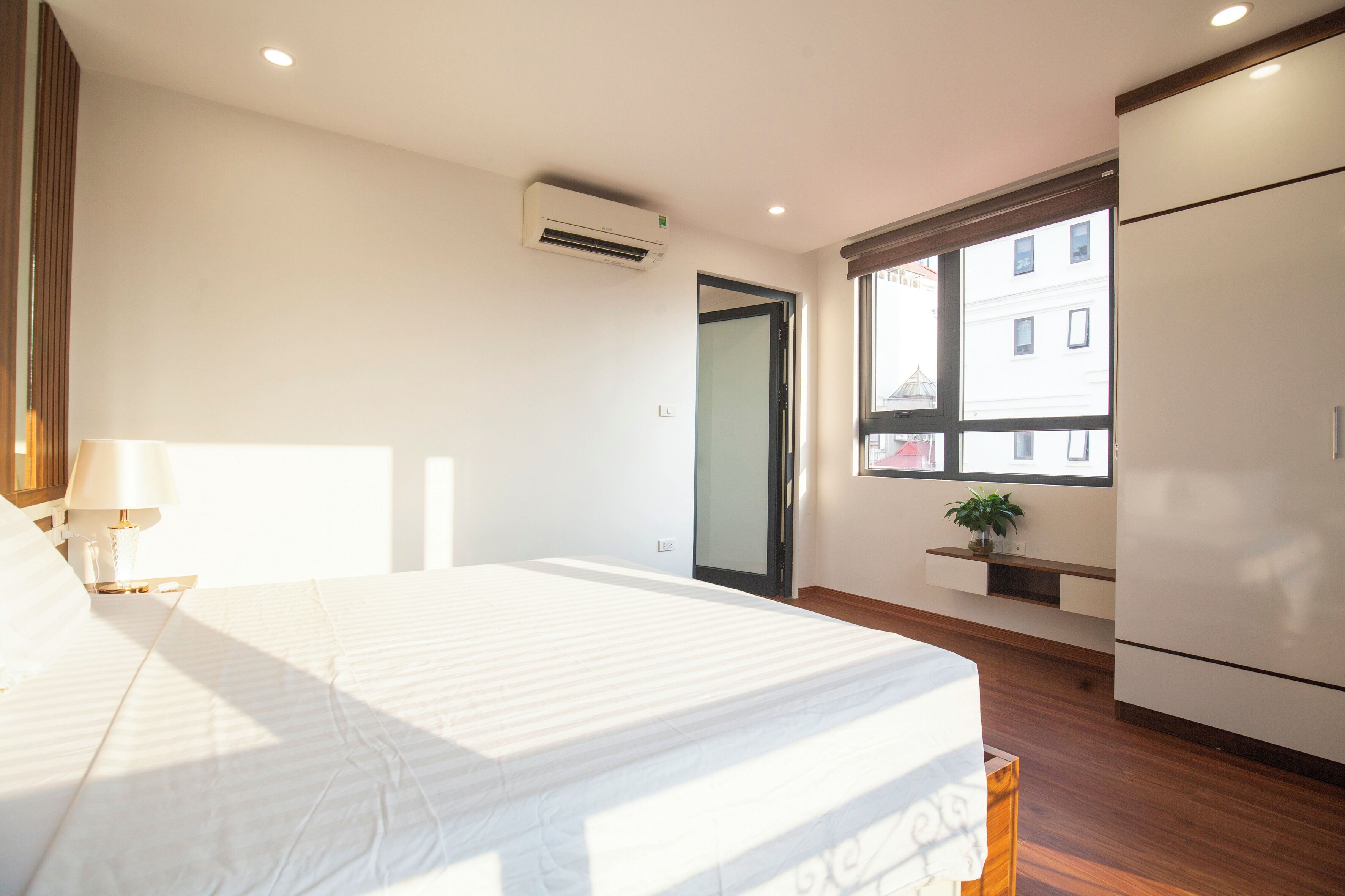 A bedroom with a bed, a nightstand, a lamp, an air conditioner, a window, and a plant.