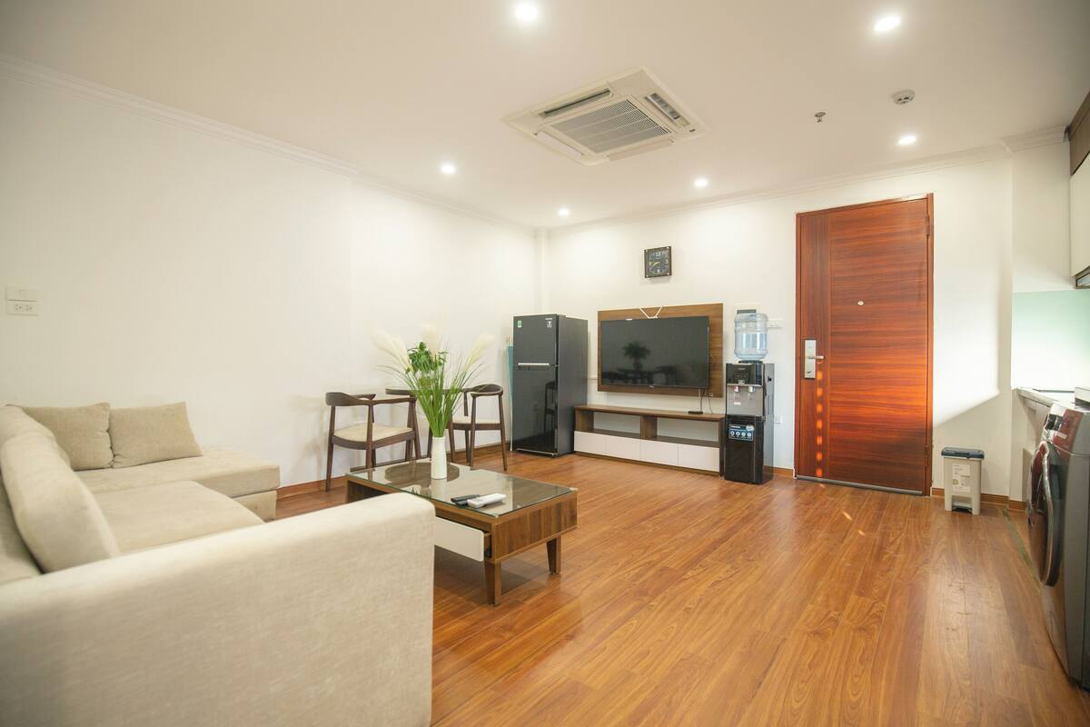 A living room with a sofa, coffee table, and a TV. There is a wooden door, a refrigerator, and a water dispenser.