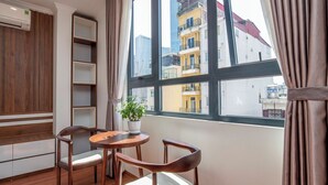 Une pièce avec un plancher en bois, une table ronde entourée de chaises, une plante en pot et une vue sur les bâtiments, visible à travers une grande fenêtre.