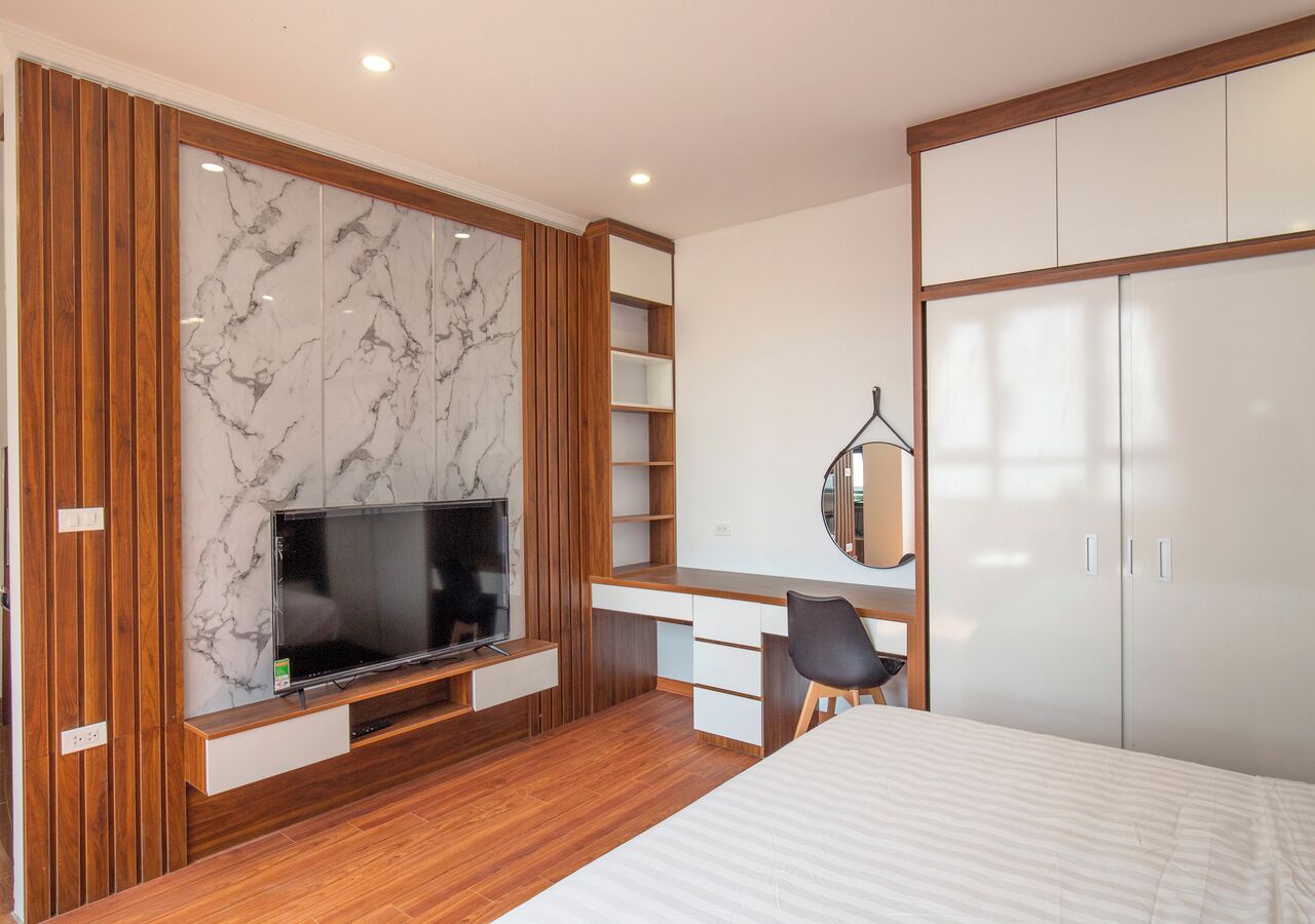 Una habitación de hotel moderna con piso de madera, una cama grande, un escritorio con una silla, un televisor en una pared de mármol y un armario.