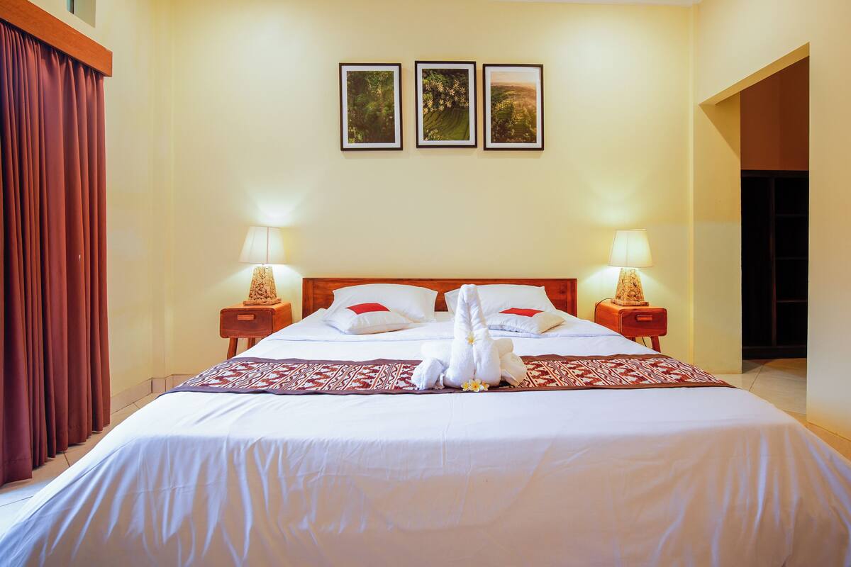 Uma cama bem arrumada com lençóis brancos e um cobertor estampado em vermelho e branco. Há duas luminárias laterais em mesas de cabeceira de madeira, e um conjunto de toalhas e uma flor sobre a cama.