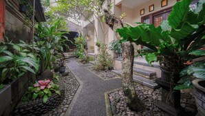 A narrow pathway flanked by lush greenery and potted plants, leading to a building with traditional architecture and a tiled roof. - Shana Homestay by EPS (Ubud)