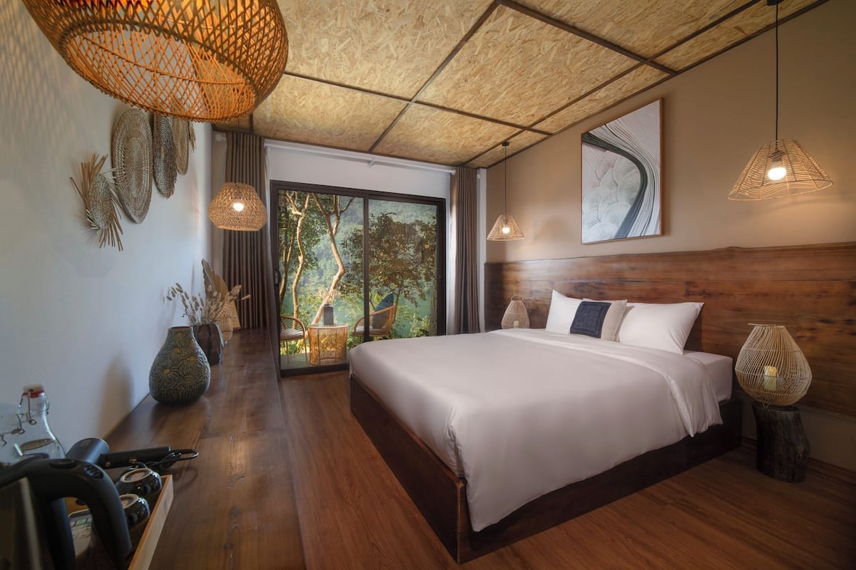 A spacious bedroom with a large bed, wooden flooring, and a view of greenery through a sliding door.