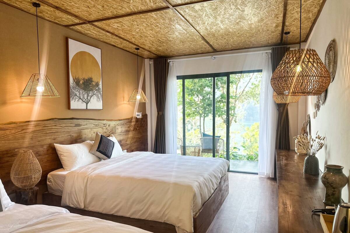 A hotel room with a large bed, two bedside tables, a wooden headboard, and a view of greenery through a glass door.