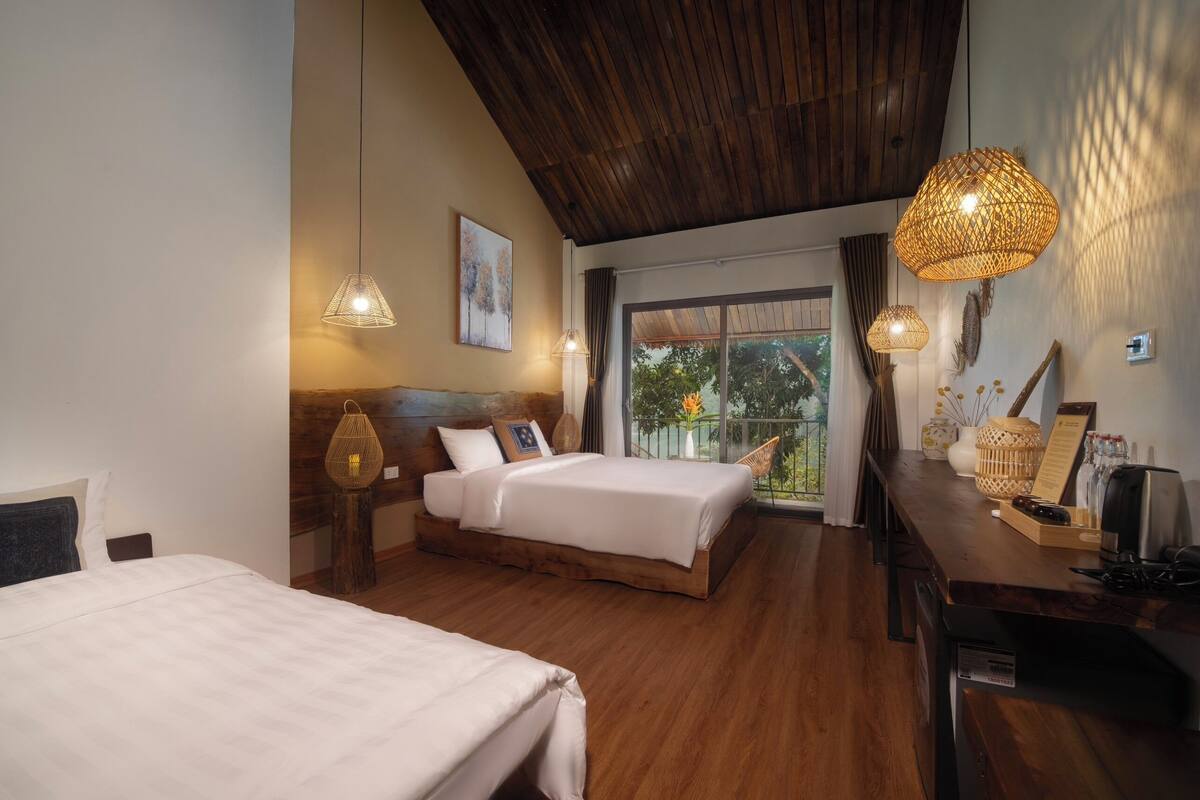 A hotel room with two beds, wooden flooring, a wooden ceiling, and a balcony with a view of trees.