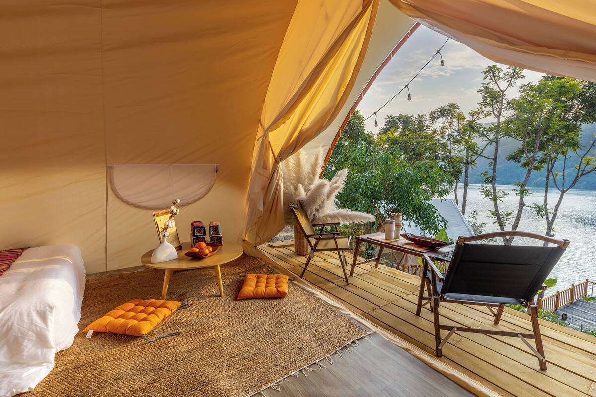 A tented room with a wooden deck, a bed, a small table with a lamp, and a view of the water.