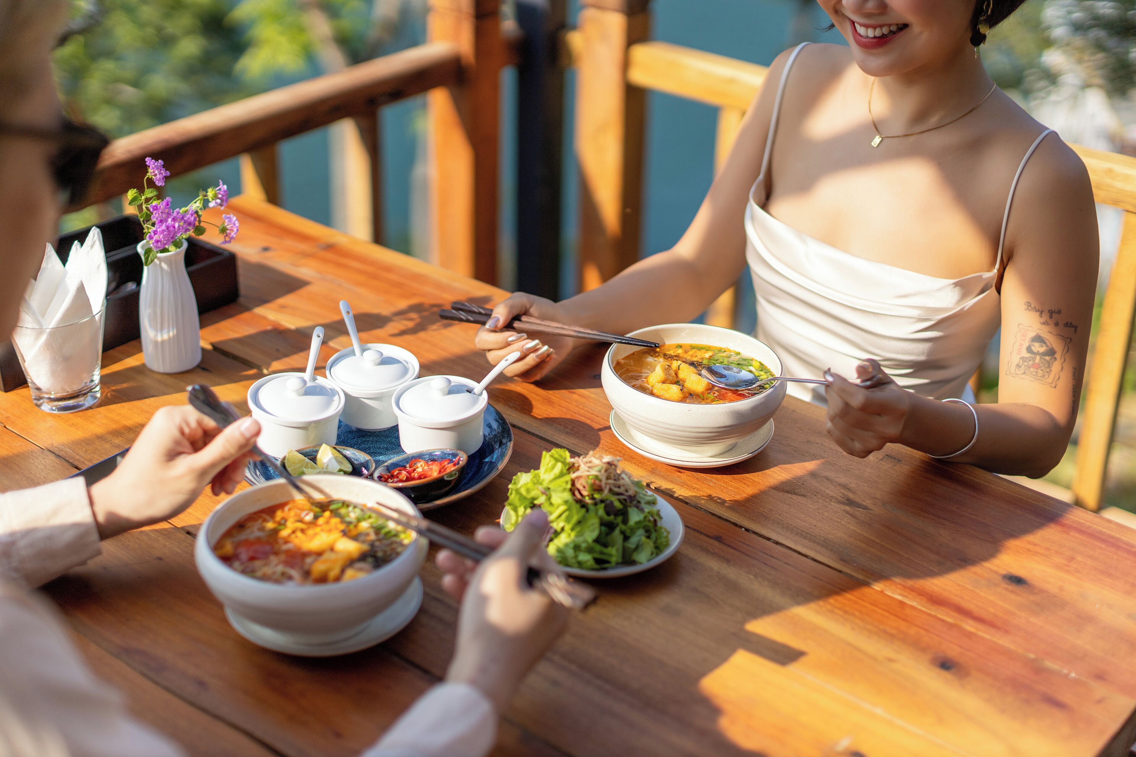 A wooden dining table with bowls of soup and salad, chopsticks, and a vase with flowers.