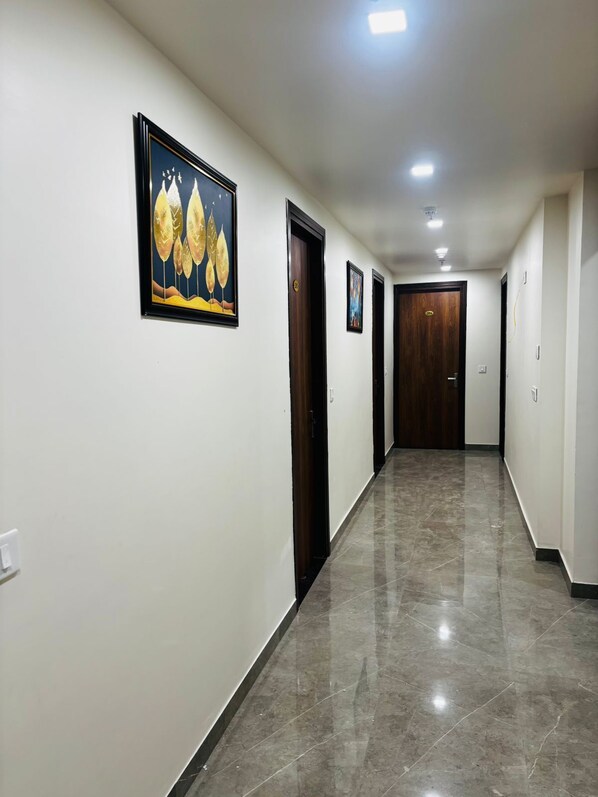 A long, narrow hallway with white walls and a polished tile floor. There are several doors along the hallway, some with visible handles. A framed picture hangs on the wall to the left. - Nirvana inn (Prayagraj)