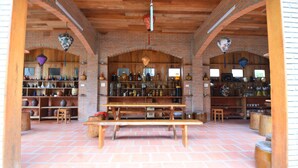 Un interior rústico con paredes de ladrillo, vigas de madera y una mesa de madera central. Estantes a ambos lados exhiben diversos objetos, y hay adornos colgantes.
